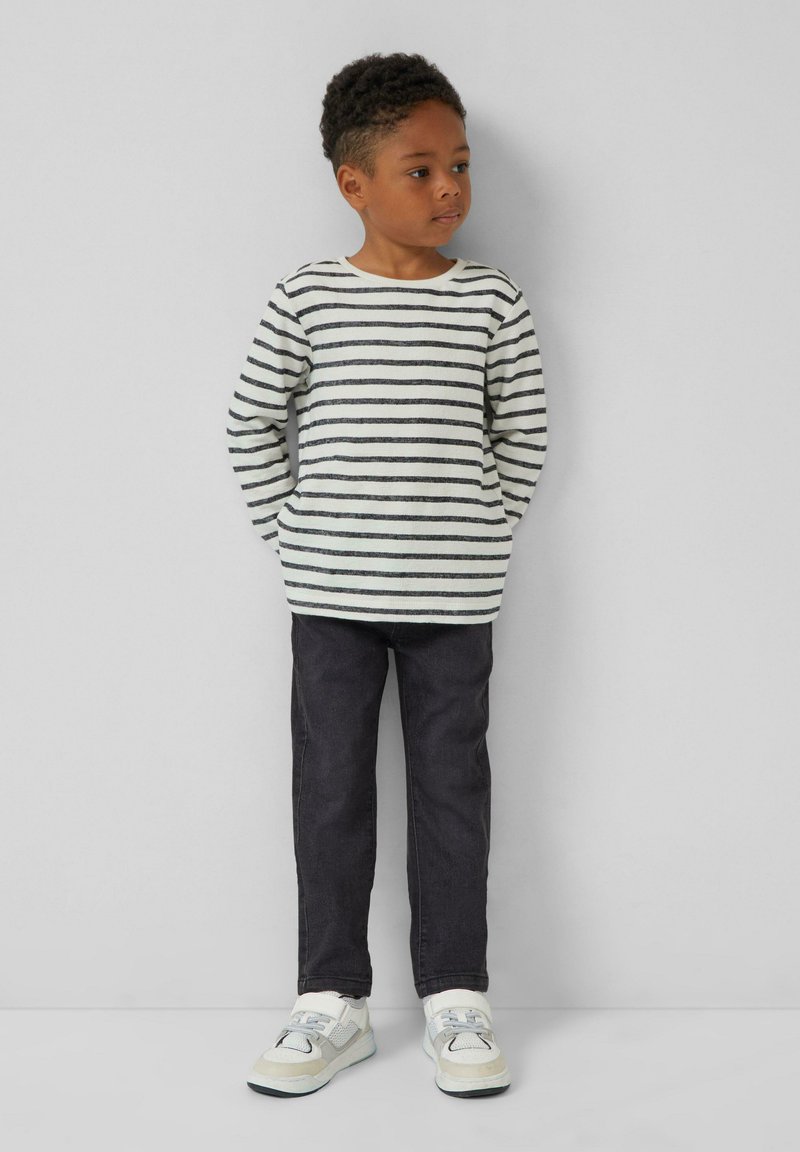Een jong kind staat tegen een effen muur en draagt een wit met zwart gestreept shirt met lange mouwen, donkere broek en witte sneakers.