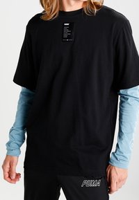 Schwarzes Baumwoll-T-Shirt mit kurzen Ärmeln, über einem hellblauen Langarmshirt getragen, mit einem Etikett auf der Brust und einem Logo am Bund.