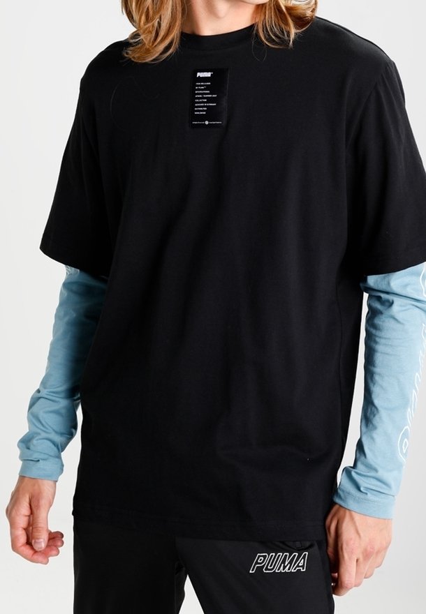 Schwarzes Baumwoll-T-Shirt mit kurzen Ärmeln, über einem hellblauen Langarmshirt getragen, mit einem Etikett auf der Brust und einem Logo am Bund.