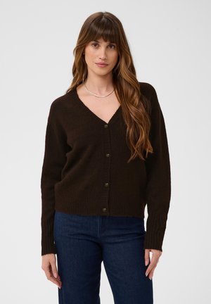 Femme aux longs cheveux bruns portant un cardigan boutonné marron foncé et un jean bleu foncé, debout devant un fond clair uni.