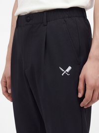 Pantalon noir avec poches latérales, taille élastique, fermeture par bouton et un motif blanc représentant un couteau et un couperet croisés sur la cuisse.