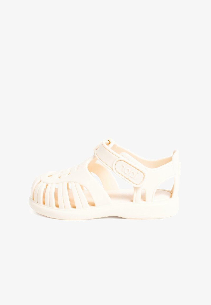 Light beige rubber sandal with a closed toe, open sides, and adjustable strap featuring a textured, cut-out design.