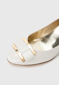 White leather shoe with a rounded toe, featuring a gold accent and a textured bow detail. The interior is gold-toned and smooth.