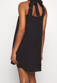Svart klänning med halterneck, spetsdetaljer på axlarna och en rosett på baksidan. Har dragkedjestängning och en utsvängd, slät textur.