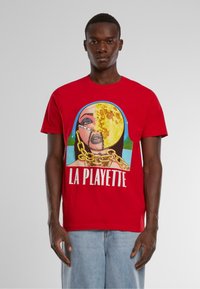 Piros pamut póló, grafikus nyomattal, amelyen egy láncos részletekkel díszített arcmás látható, alján pedig fehér színnel a "LA PLAYETTE" felirat olvasható.