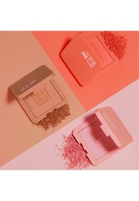 Deux compacts de blush rectangulaires : l'un beige avec une finition mate, l'autre rose avec une texture scintillante, tous deux entourés de poudre libre.