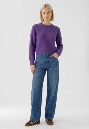 Femme blonde debout avec une main dans la poche, portant un pull violet duveteux, un jean bleu à jambes larges et des baskets marron avec des bandes blanches.