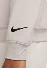 Svetlo šedý rukáv mikiny vyrobený z jemného materiálu, ktorý obsahuje čierne logo Nike swoosh a ribbové manžety s textúrovaným povrchom.