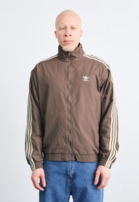 Brun zip-up jakke med høy krage, prydet med hvite striper på ermene og et brodert logo på brystet. Lett materiale.