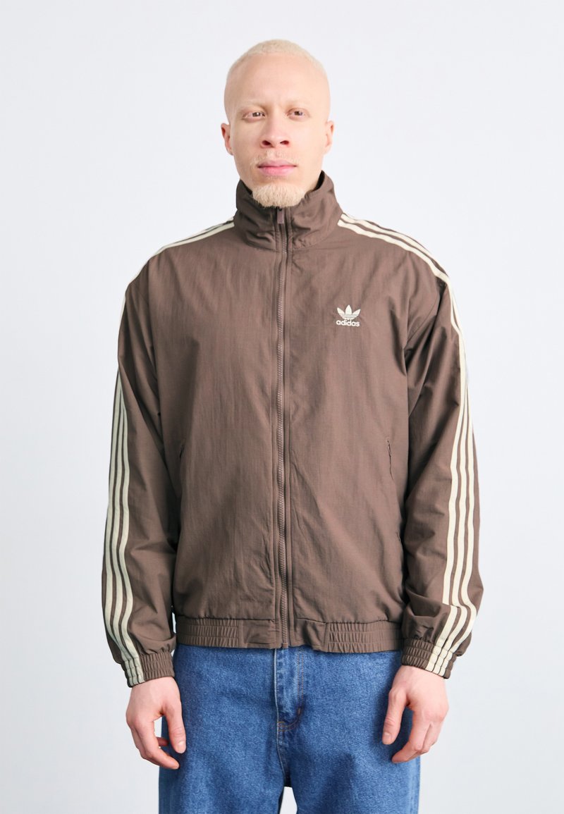 Brun zip-up jakke med høy krage, prydet med hvite striper på ermene og et brodert logo på brystet. Lett materiale.