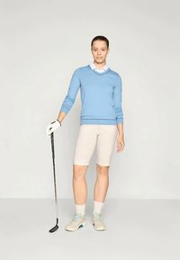 Mulher em pé segurando um taco de golfe com a mão enluvada, vestindo um suéter azul, camisa branca de colarinho, calções bege e sapatos de golfe de cor clara.