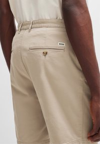 Bermudas beige de un tejido ligero, con un bolsillo trasero con cierre de botón y una pequeña etiqueta blanca "BOSS" en la cinturilla.
