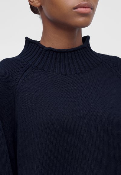 Gros plan d'une personne portant un pull en maille bleu marine foncé avec un col montant côtelé.