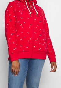 Czerwona bluza z kapturem na zamek błyskawiczny, z kapturem wyposażonym w sznurek, ozdobiona kwiatowym wzorem w kolorach niebieskim i białym. Materiał wydaje się miękki i elastyczny, z prążkowanymi mankietami.