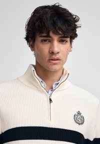 Junger Mann mit dunklem lockigem Haar, der einen cremefarbenen Strickzippullover mit schwarzem Streifen und gesticktem Logo auf der Brust trägt.