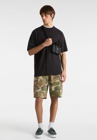 Černé oversized tričko, maskáčové cargo šortky, zelené sneakers a malá černá crossbody taška. Jednoduché pozadí; žádné další detaily.