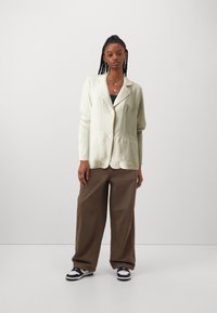 Light beige button-up blazer with a textured finish, paired with loose brown trousers and black and white sneakers. Simple design, relaxed fit.