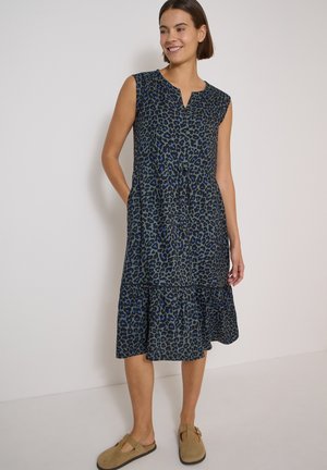 Donna che indossa un abito midi senza maniche con stampa leopardata blu scuro e nero, con orlo arricciato e scarpe slip-on color cuoio, in piedi contro un muro bianco.