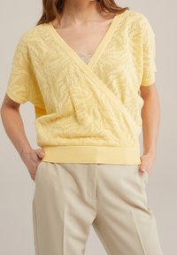 Gul, texturerad stickad topp med V-ringning och korta ärmar, med omslagsdesign och ribbad nederkant. Kombinerad med ljusbeige byxor.