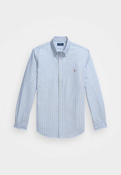 Lys blå knapp-up skjorte med tynne hvite striper, laget av bomull. Har en sprettkrage og en liten brodert logo på brystet.