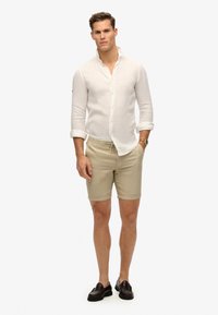 Camisa blanca de lino de botones, pantalones cortos beige y mocasines oscuros. La camisa tiene las mangas enrolladas; los pantalones cortos cuentan con una cinturilla elástica y bolsillos.