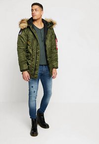 Olivgrüne Puffjacke mit gefütterter Kapuze, Seitentaschen, schwarzem Innenfutter, kombiniert mit blauen, ausgewaschenen Jeans und schwarzen Schnürstiefeln.