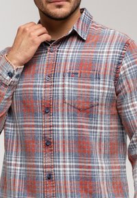 Mężczyzna poprawiający kołnierzyk niebieskiej, czerwonej i białej koszuli w kratę z kieszonką na piersi oraz subtelnym haftowanym logo.