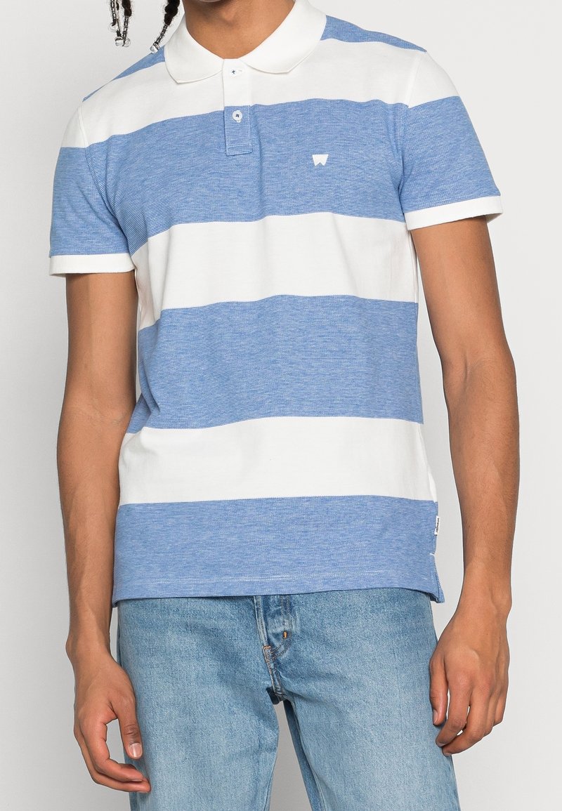 Homme portant un polo à manches courtes rayé bleu et blanc, avec un jean bleu clair, se tenant devant un fond uni.