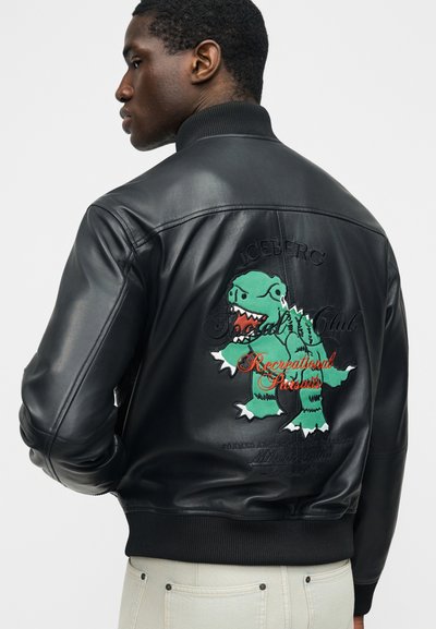 Άνδρας με μαύρο δερμάτινο μπουφάν με πράσινο μπάλωμα δεινόσαυρου και κόκκινο κείμενο "Recreational Pursuits" στην πλάτη, ορατό άσπρο παντελόνι.