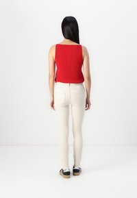 Femme aux longs cheveux noirs portant un haut rouge sans manches, un pantalon blanc ajusté et des baskets noires et blanches, debout face à un mur blanc.