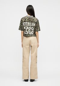 Γυναίκα που στέκεται με την πλάτη γυρισμένη φορώντας κοντομάνικο πουκάμισο με μοτίβο λεοπάρδαλης και κείμενο «ICECREAM IC» καθώς και μπεζ παντελόνι cargo με μαύρα παπούτσια.