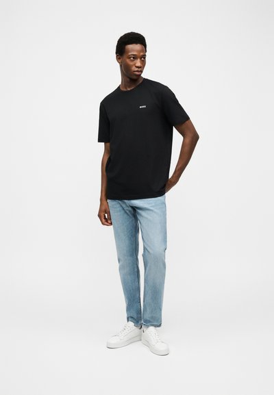 Jeune homme debout, main dans la poche, portant un T-shirt noir BOSS, un jean bleu clair et des baskets blanches sur fond gris uni.