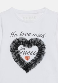 Λευκό πουκάμισο με γκλίτερ κείμενο «Ερωτευμένη με τη Guess», με μια μαύ ρη καρδιά με βολάν και μια μικρή κόκκινη καρδιά με γκλίτερ κάτω από το κείμενο.
