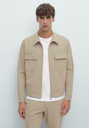 Veste zippée beige avec un col, comportant deux poches avant. La veste a une texture lisse, assortie à un t-shirt blanc en dessous.