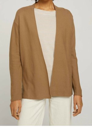 Personne portant un cardigan marron clair ouvert devant sur une chemise rayée blanche et beige avec un pantalon blanc, debout devant un fond uni.