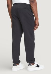 Schwarze Sweatpants aus weichem Stoff, mit elastischem Bund, schmal zulaufendem Bein und einer einzigen Gesäßtasche. Tragen mit weißen Turnschuhen.