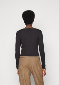 Person med kort lockigt hår sedd bakifrån, iförd en svart långärmad topp med ett leopardmönstrat halslinning och beige byxor.