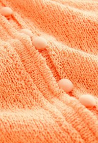 Vêtement tricoté en orange vif avec un tissu texturé. Présente des boutons ronds et texturés sur le devant, mettant en valeur le détail de design.