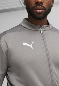 Graue Zip-Jacke aus strukturiertem Stoff, mit hohem Kragen und weißem Puma-Logo auf der linken Brust, sowie kontrastierendes Schulterpartiendesign.