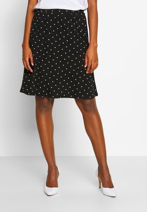 Jupe noire à pois blancs, avec une taille élastique et un design fluide qui tombe juste au-dessus du genou. Assortie à des talons blancs.
