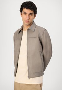 Jaqueta de couro cinza com colarinho, fecho frontal e bolsos laterais. Apresenta uma textura suave e um design minimalista, combinada com uma camisa bege.