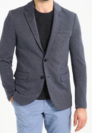 Mies pukeutuneena tummanharmaaseen teksturoituun bleiseriin, mustaan paitaan ja vaalean sinisiin housuihin, seisomassa yhdellä kädellä taskussa.