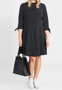 Černé šaty s bílými puntíky, tříčtvrteční rukávy s vázacími detaily, doplněné černou taškou přes rameno a bílými teniskami.
