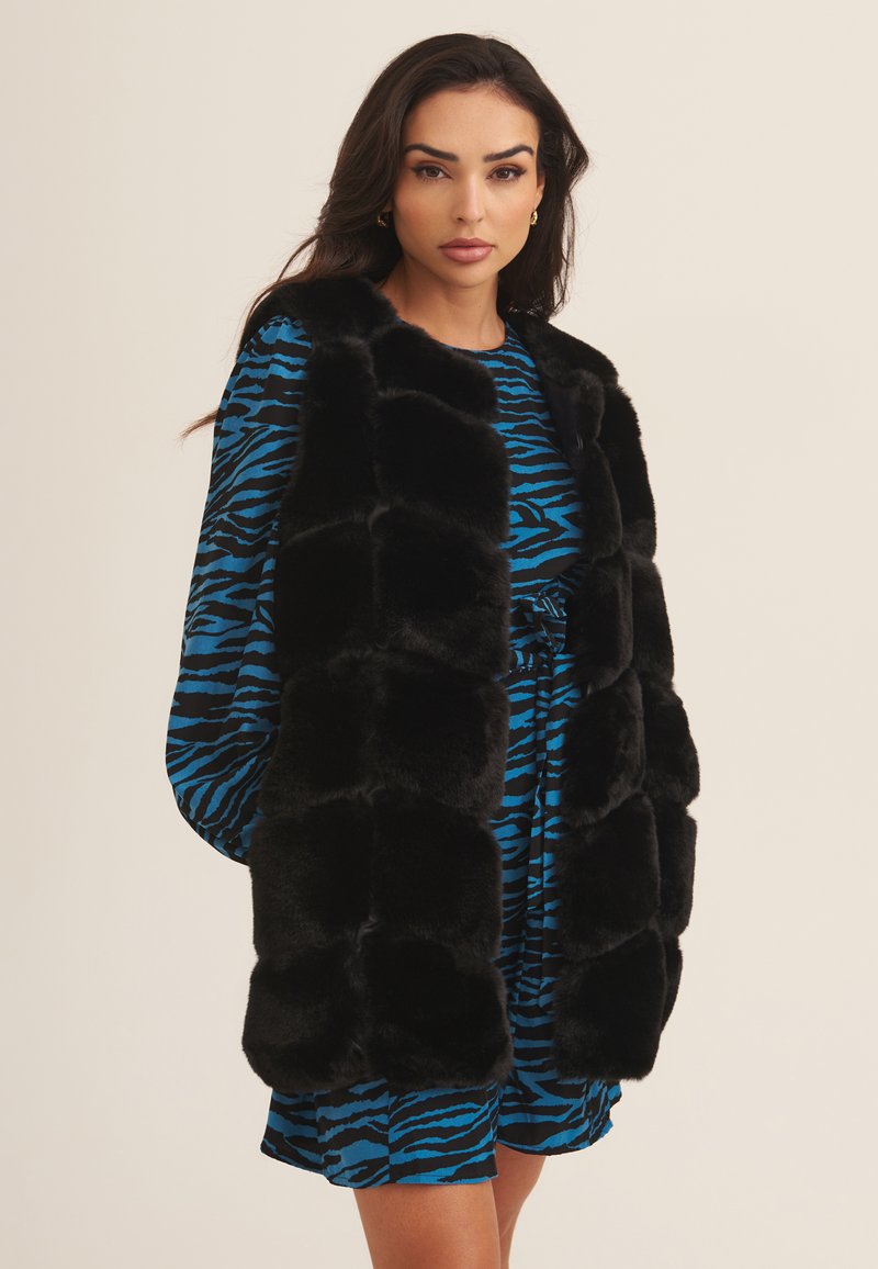 Black faux fur vest with plush texture, featuring a quilted design. Worn over a blue and black striped patterned dress.