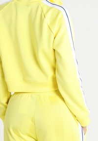 Gul kortärmad sweatshirt med vita sidostreck; texturerad tyg; ribbade mudd; parat med matchande gula byxor som visar vertikala mönster.