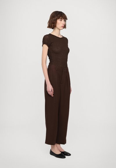 Brown textured knit top with short sleeves, paired with high-waisted brown trousers. Model wears black flat shoes. Plain background.