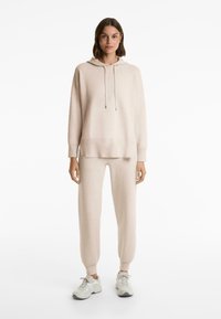 Femme debout portant un hoodie beige et un pantalon de jogging assorti avec des baskets blanches sur un fond blanc.
