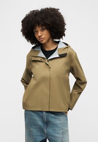 Jauna moteris su garbanotais plaukais dėvi rudos spalvos neperšlampamą striukę ant juodo viršaus ir laisvų mėlynų džinsų, stovinti priešais paprastą foną.