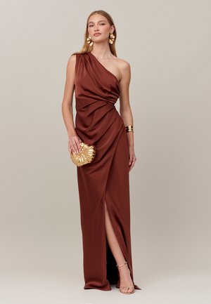 Femme en robe satinée rouille à une épaule, tenant une pochette dorée en forme de coquillage, portant des boucles d’oreilles, un bracelet et des talons à brides dorés.