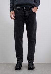 Hombre vestido con pantalones vaqueros negros y zapatos de cuero negros con cordones, de pie contra un fondo gris claro liso.